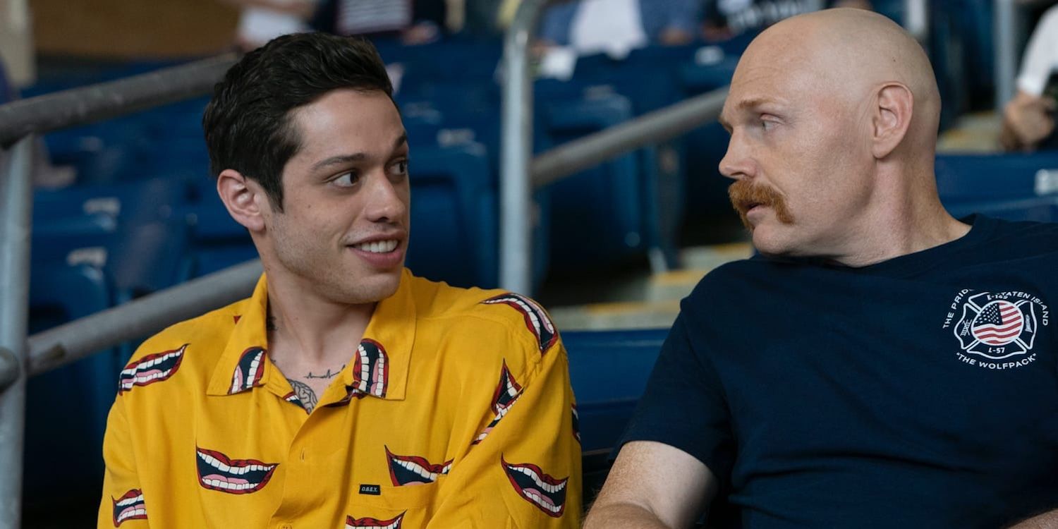 Pete Davidson and Bill Burr sitting in chairs at a baseball stadium, looking at each other, in The King of Staten Island