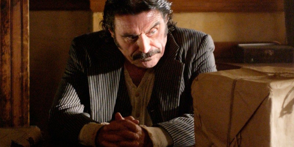 Ian McShane sitting behind a desk with a wrapped box in front of him