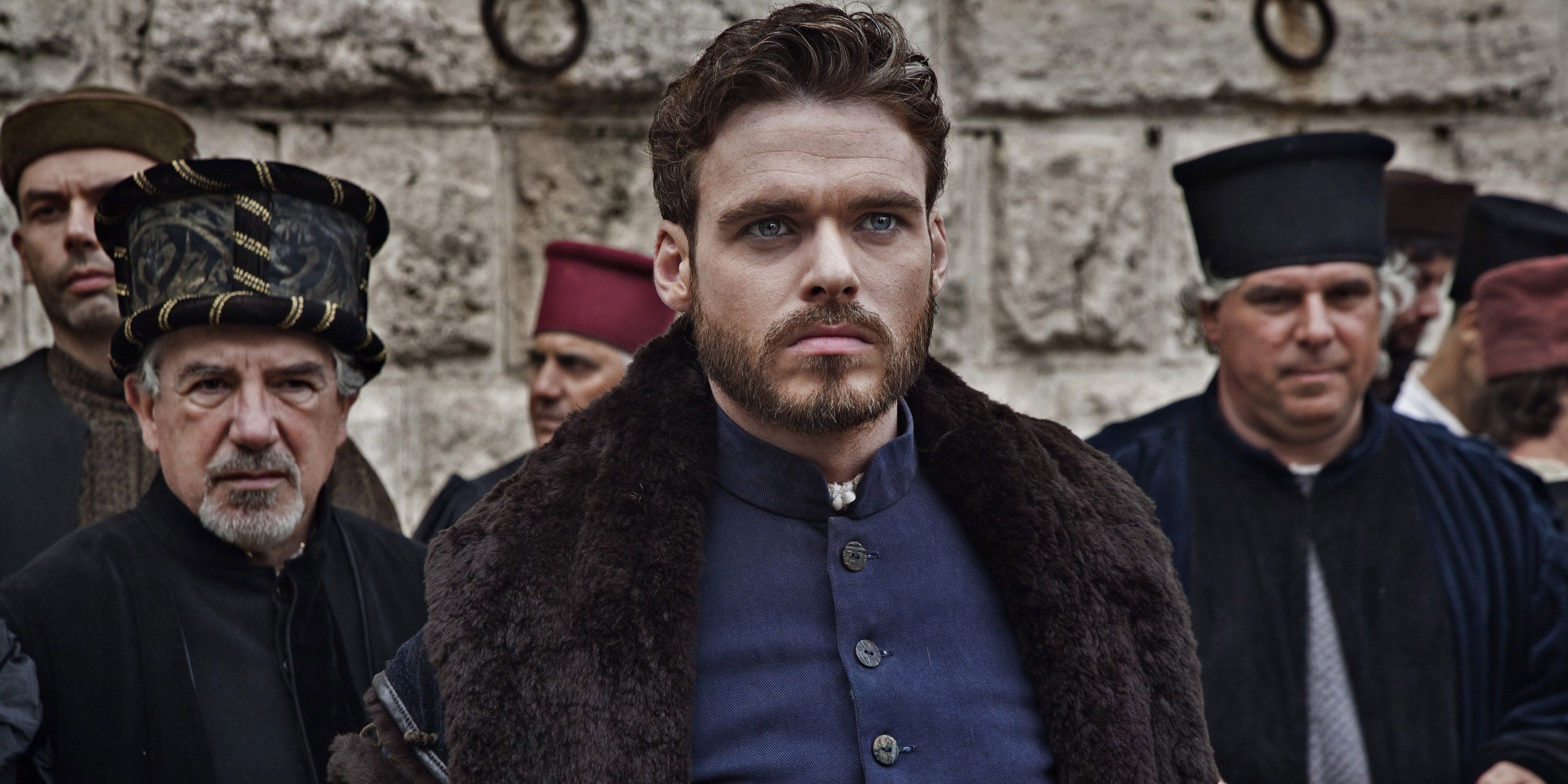 Richard Madden in a blue suit and fur surrounded by people in Medici.
