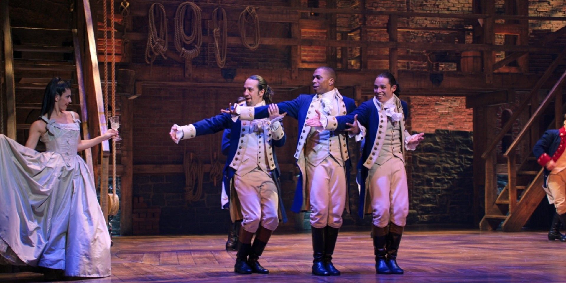 Alexander Hamilton and his friends gesture to a woman coming down the stairs in Hamilton