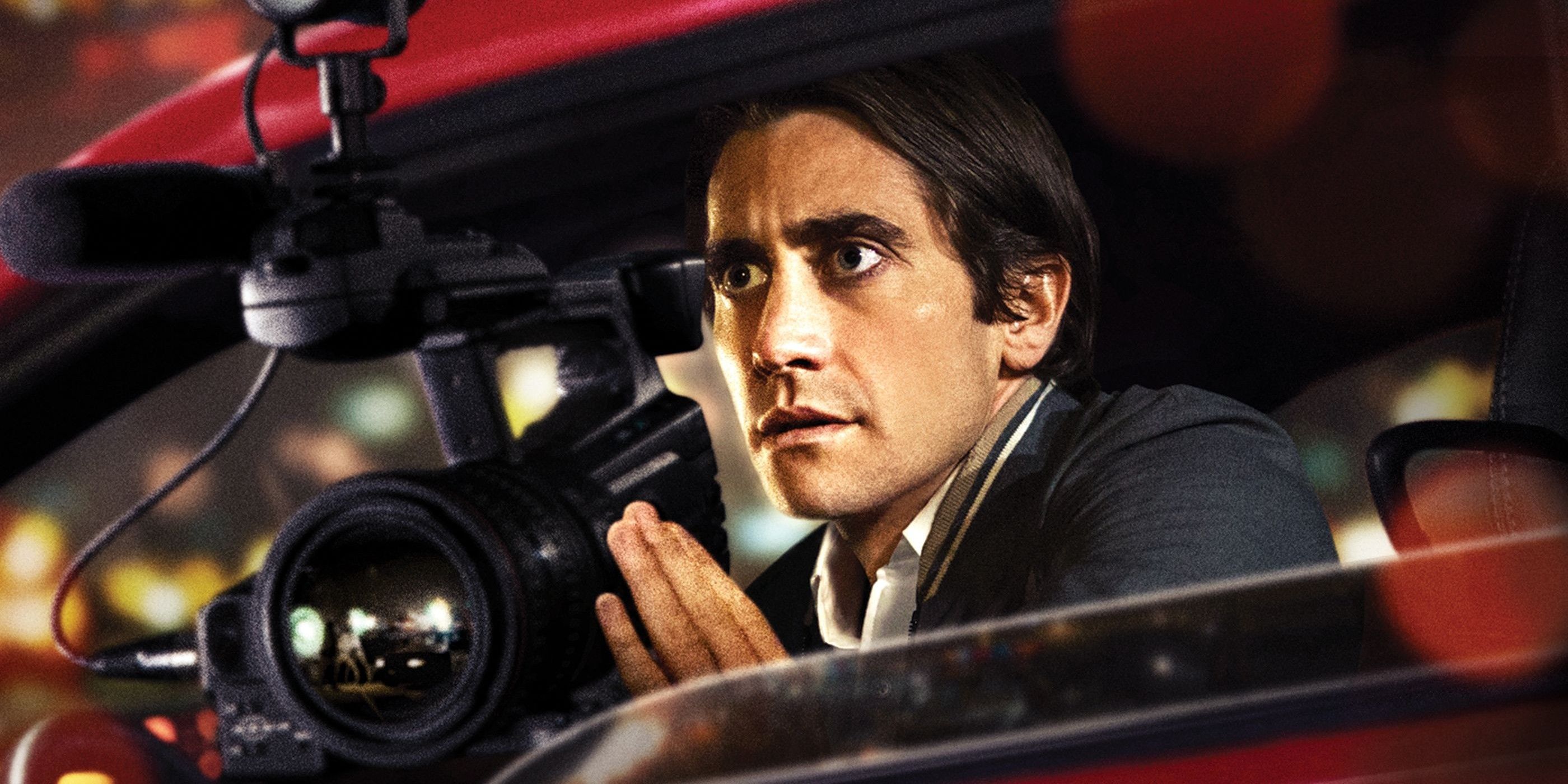 Lou Bloom looking out the window of his car while holding a camera in Nightcrawler Banner