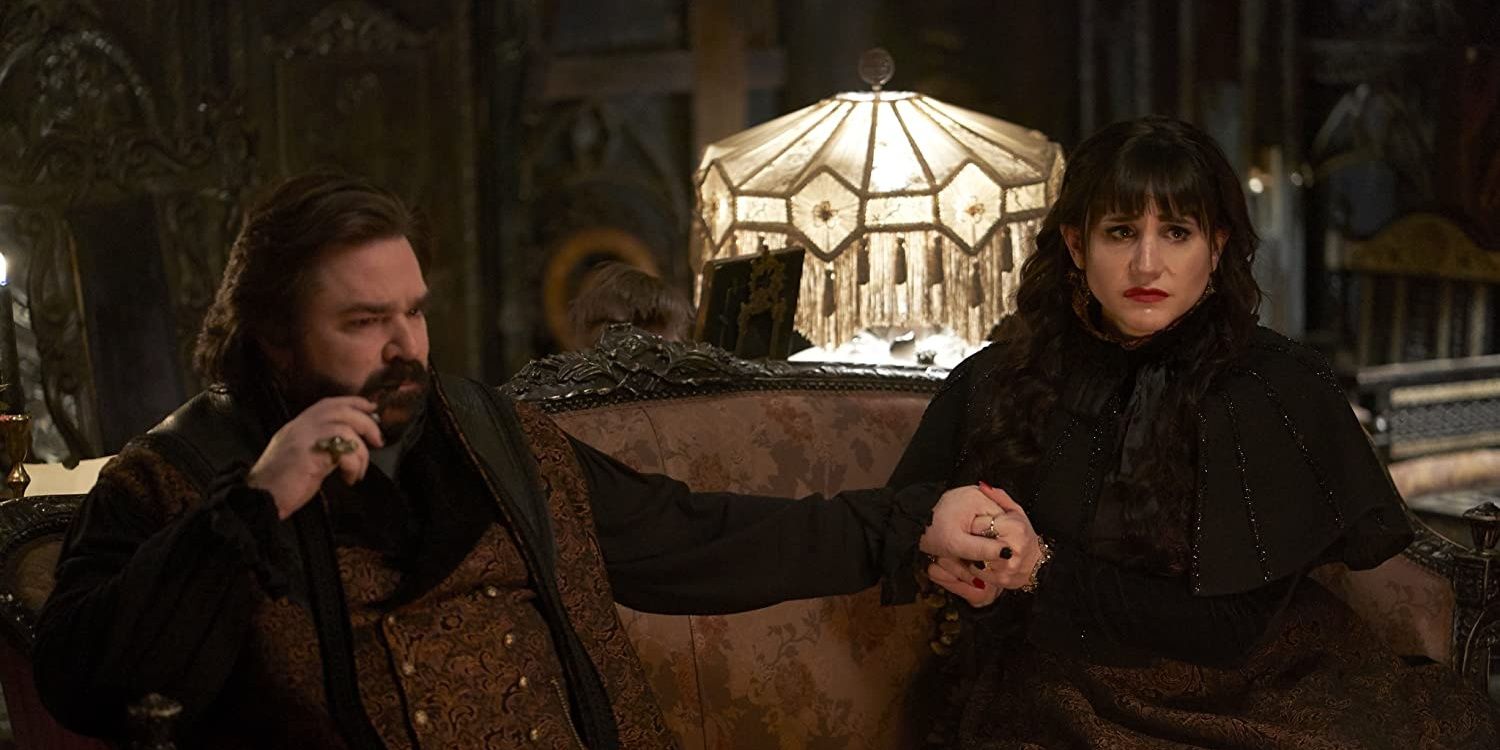 Laszlo holding Nadja's hand as they sit on a couch in What We Do In The Shadows.