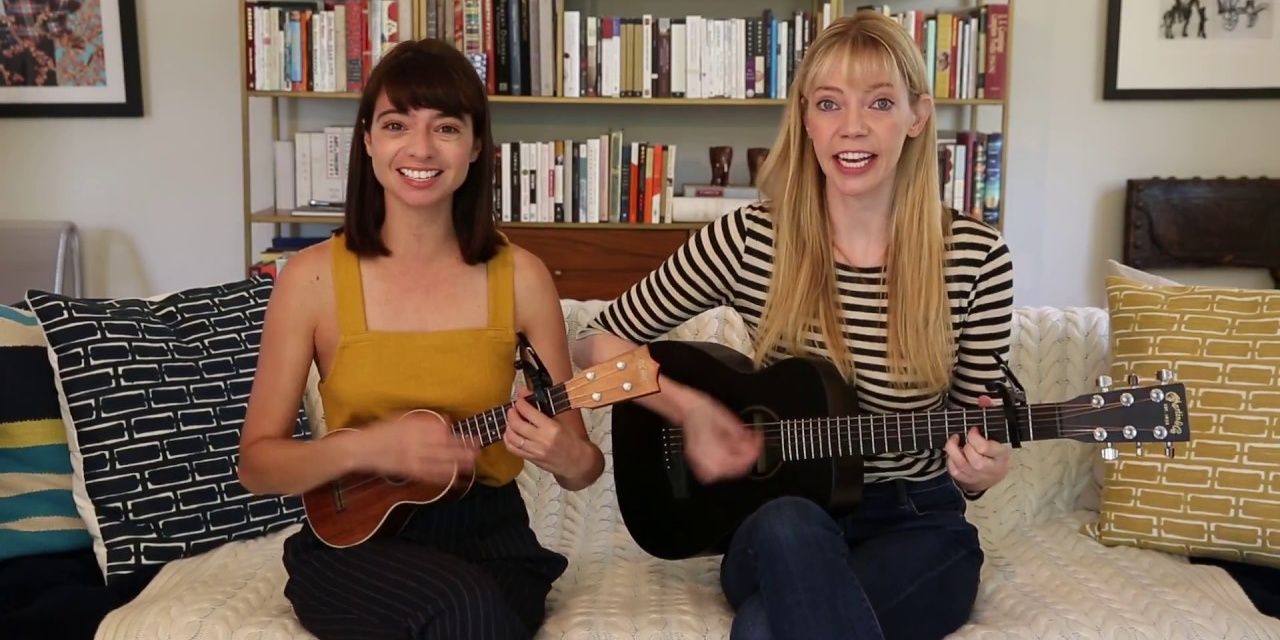 Garfunkel and Oates sitting side by side playing guitar