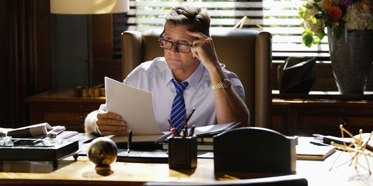 Blake Carrington from Dynasty sitting at his desk looking at papers with glasses on.