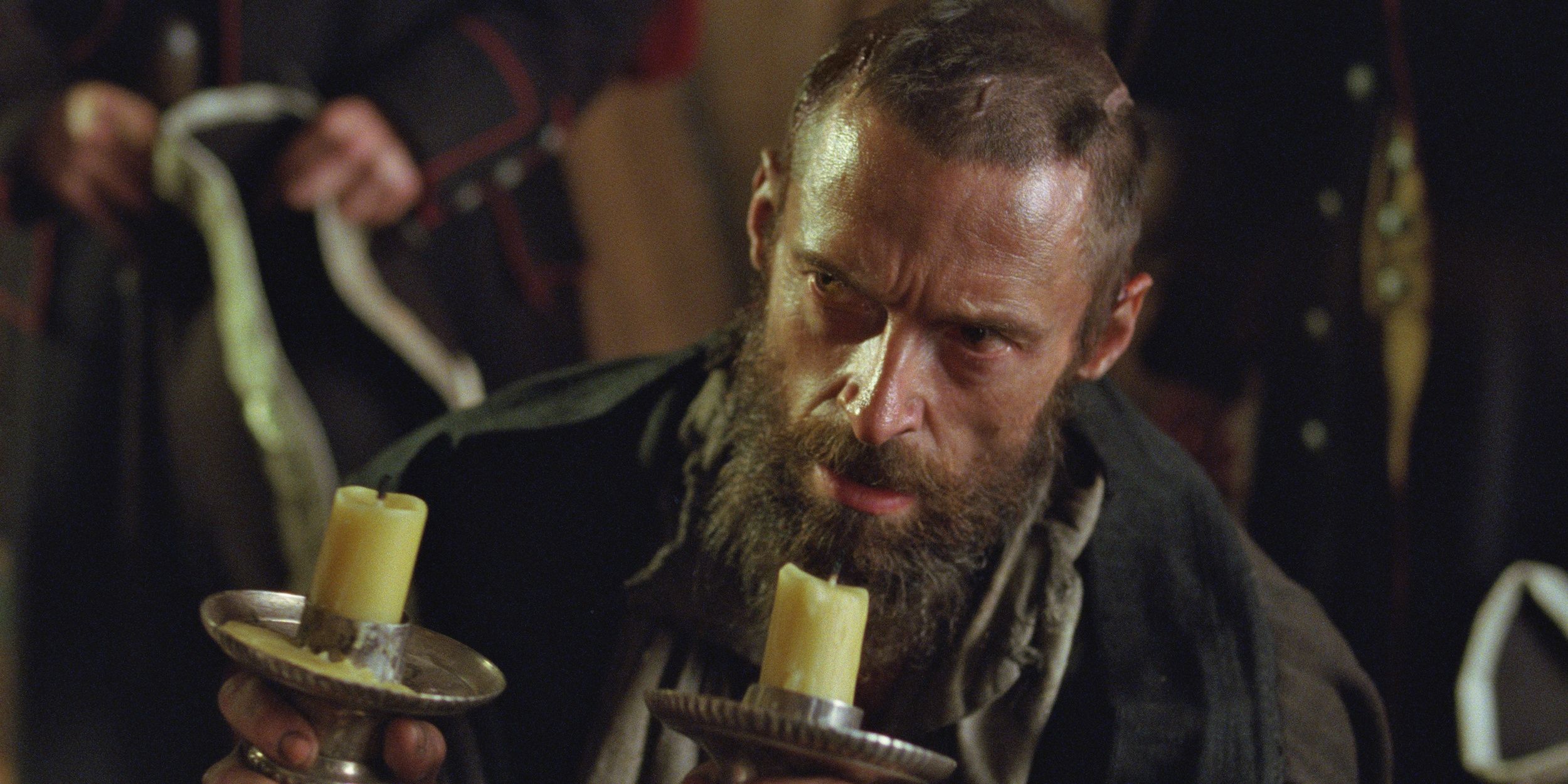 Hugh Jackman in Les Misérables looking scruffy with shaved head and beard holding two candles 