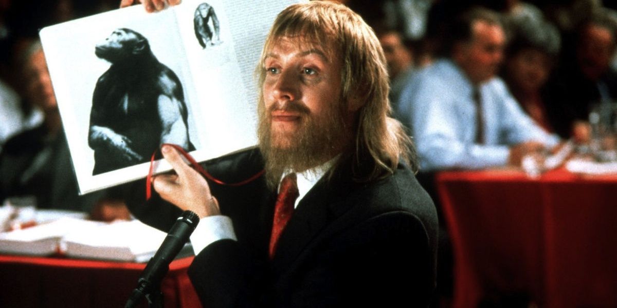 Rhys Ifans pointing to an image of an ape in a book in a still from Human Nature