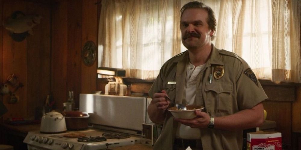Jim Hopper from Stranger Things leans against the sink while eating cereal and smiling