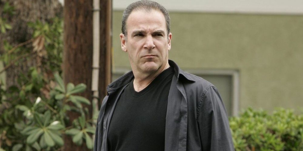 Mandy Patinkin scowls in front of some bushes.