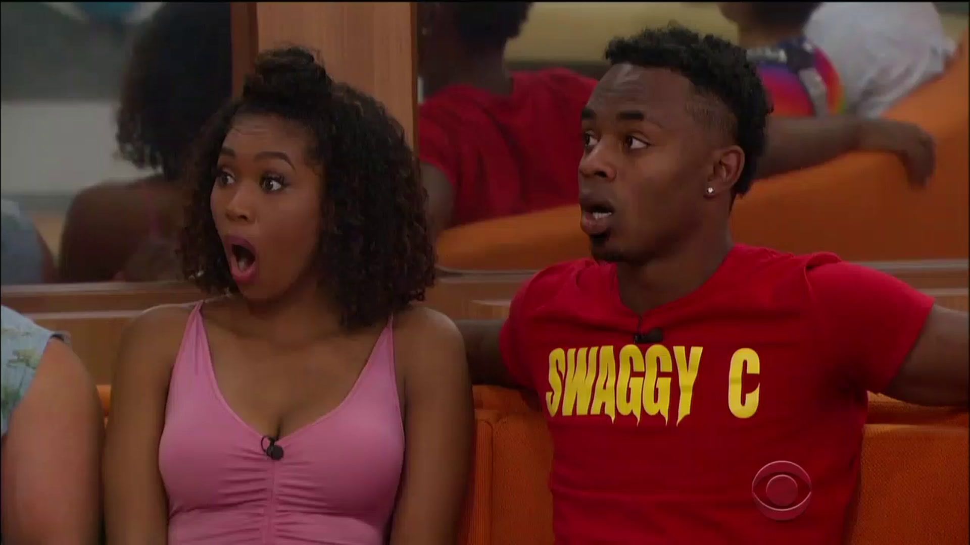 Swaggy C sitting with Bayleigh on the couch on eviction night in Big Brother, both with their mouths agape in shock. She is wearing a pink tank top and he a red T-shirt with the words &quot;Swaggy C&quot; written across the middle in yellow