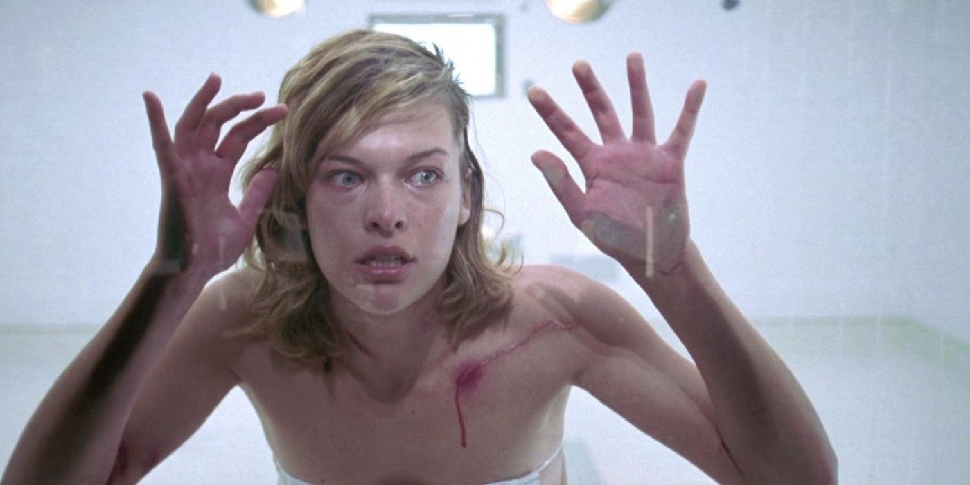 Milla Jovovich como Alice parecendo chocada com as mãos e o rosto contra o vidro no primeiro filme de Resident Evil
