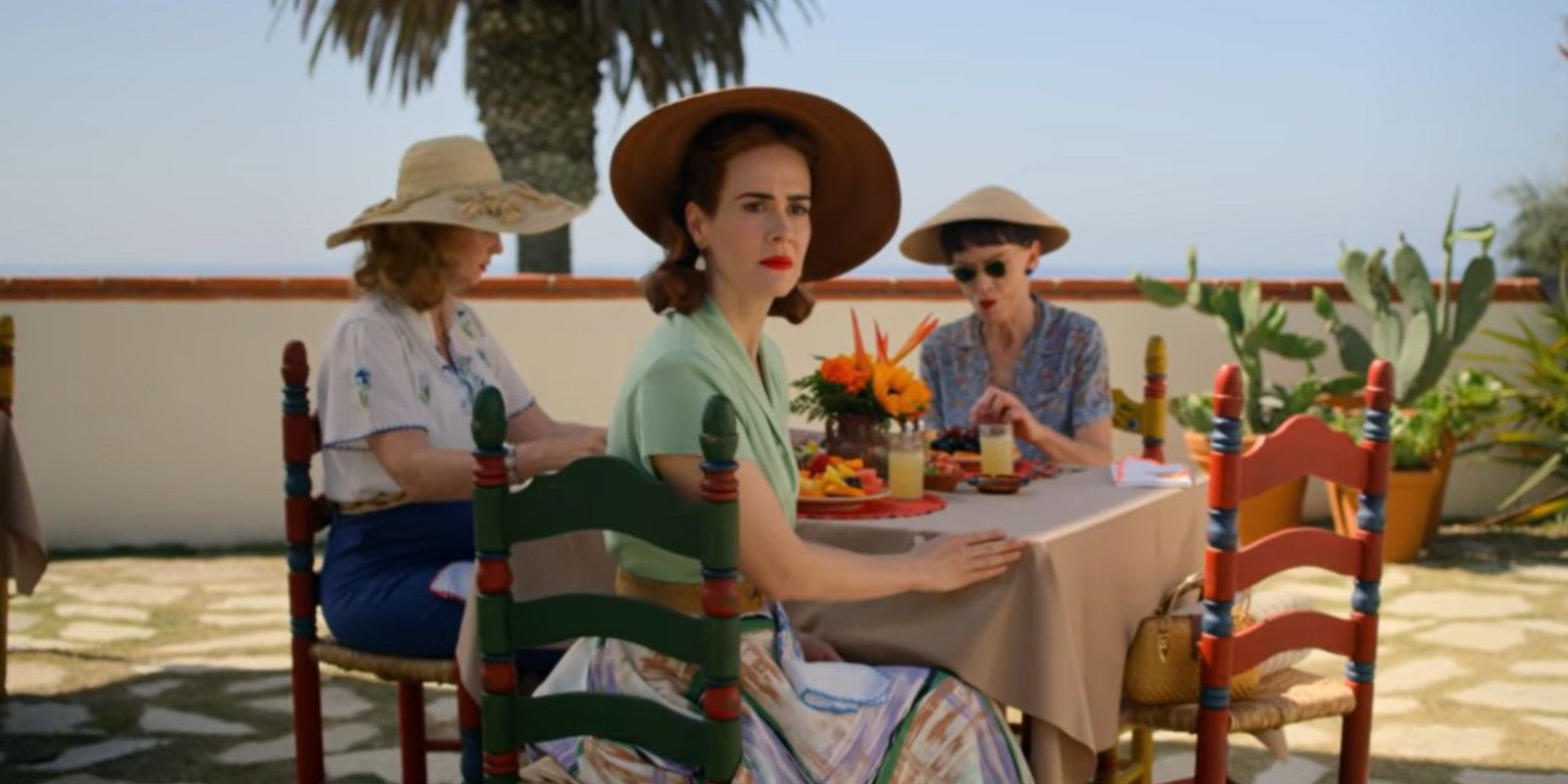Ratched (Sarah Paulson) sitting outdoors at a table turning back and looking concerned in Ratched.