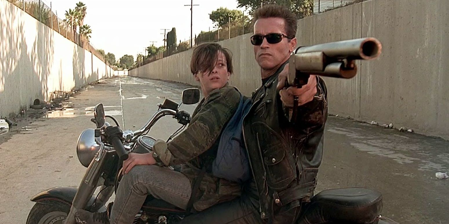 T-800 and John Connor sit on a motorcycle, T-800 pointing a shotgun.