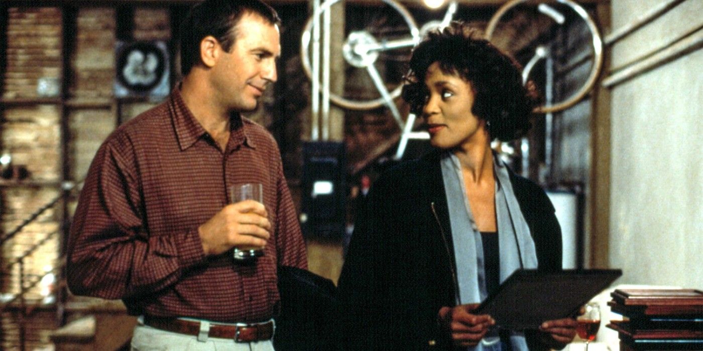 Whitney Houston as Rachel holding a folder and Kevin Costner as Frank holding a glass of beverage as they look at each other in The Bodyguard