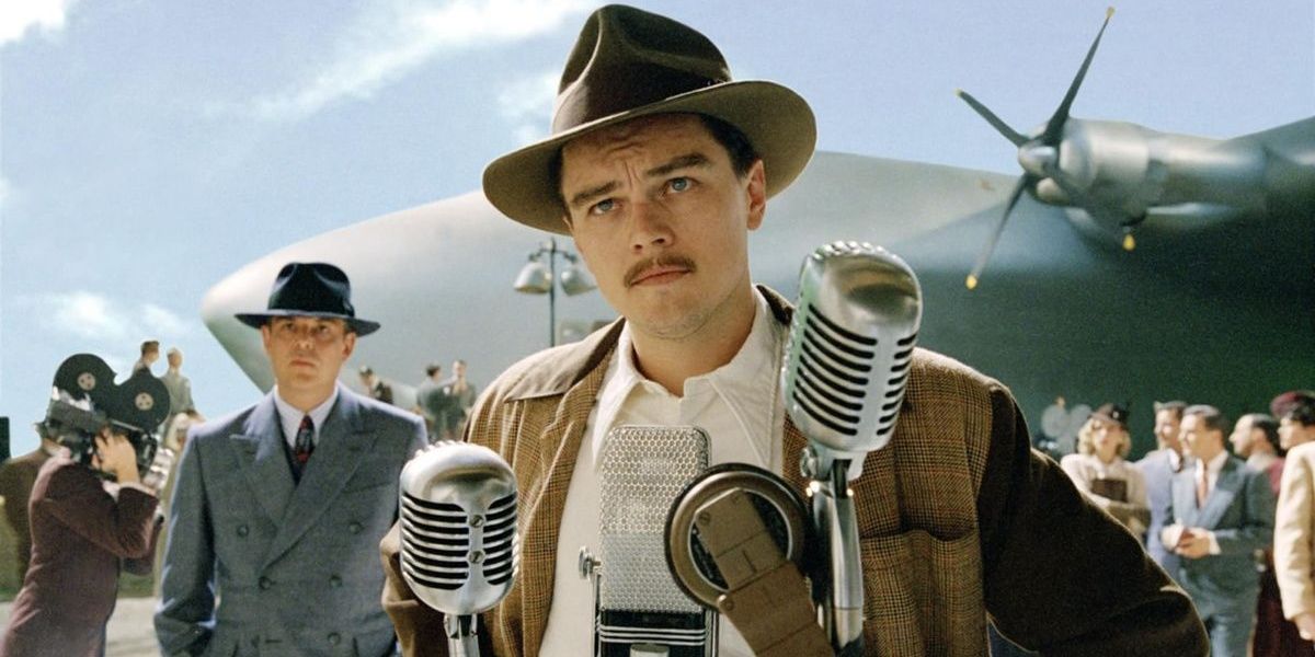 Leonardo Dicaprio in The Aviator standing at a microphone and in front of an airplane wearing a hat. 