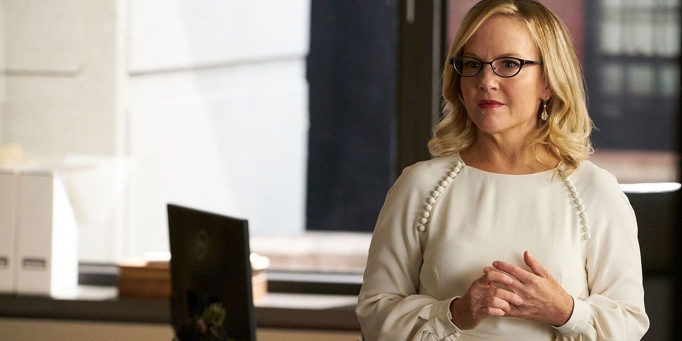 Shelia from Suits with her hands together, wearing a white sweater and dark glasses.