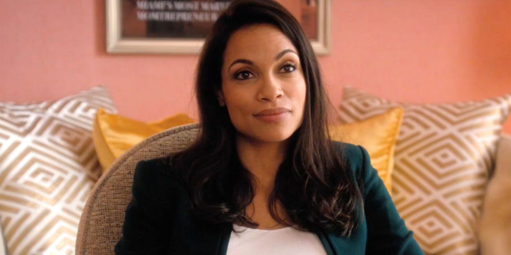 Rosario Dawson sitting on a couch with a pink wall in the background