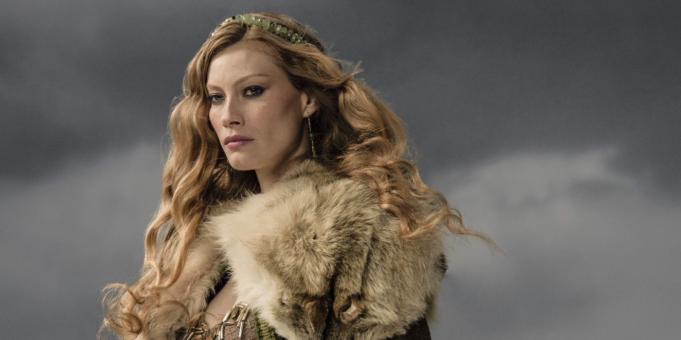 Aslaug in Vikings standing in fur with flowing hair and clouds in the background, with an ominous look on her face.