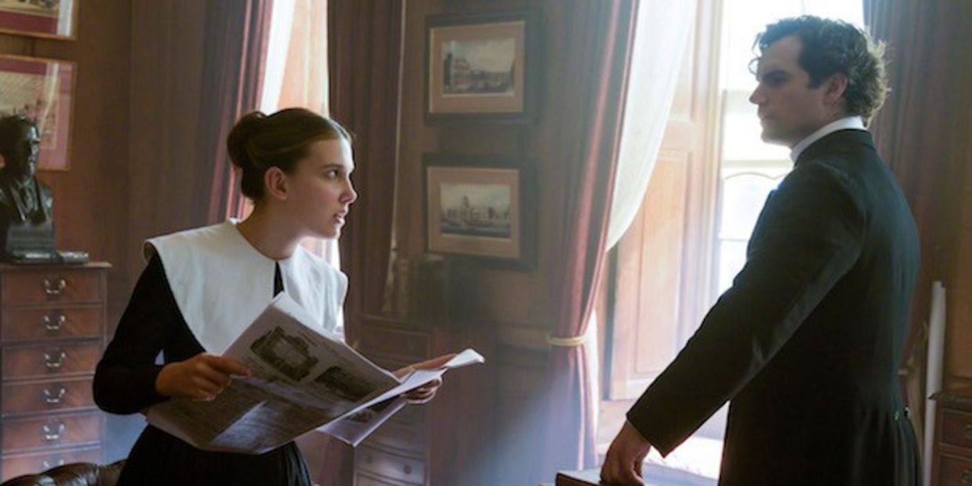 Enola holds an open newspaper and talks to Sherlock