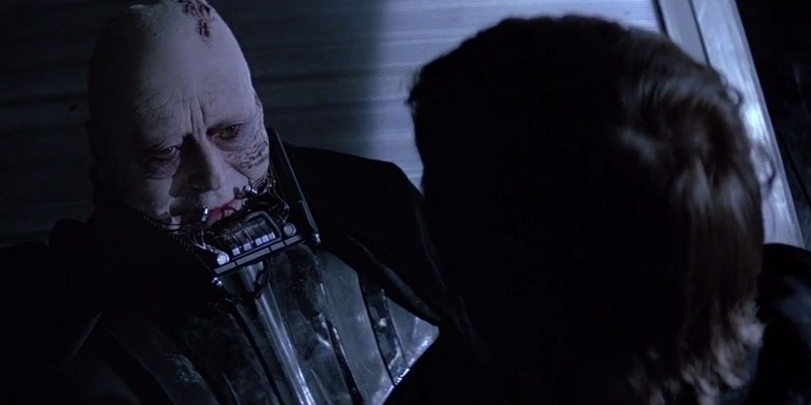 Luke sits in front of Darth Vader, whose mask is off, showing his face. 