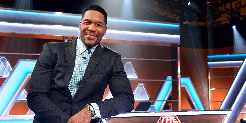 Michael Strahan leaning against a desk and smiling for the camera.