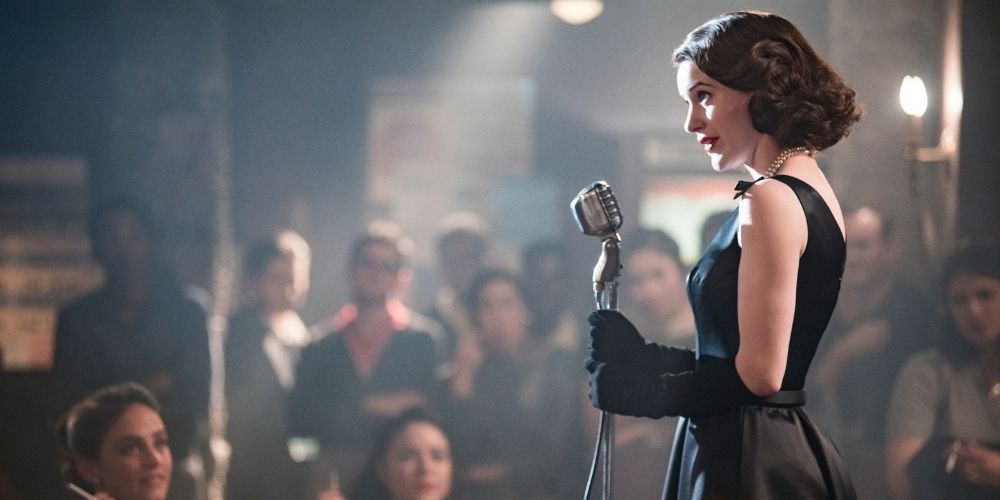 Midge talking into a microphone on stage talking to the audience in The Marvelous Mrs. Maisel. 