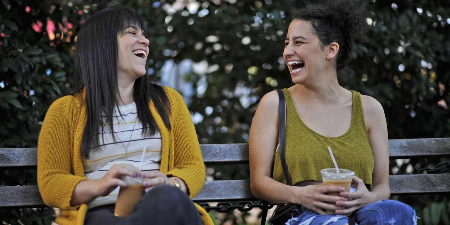 Ilana and Abbi sitting on a bench and laughing in Broad City