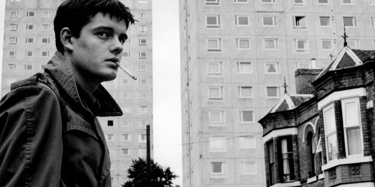 Ian Curtis stands in front of a grey building from Control