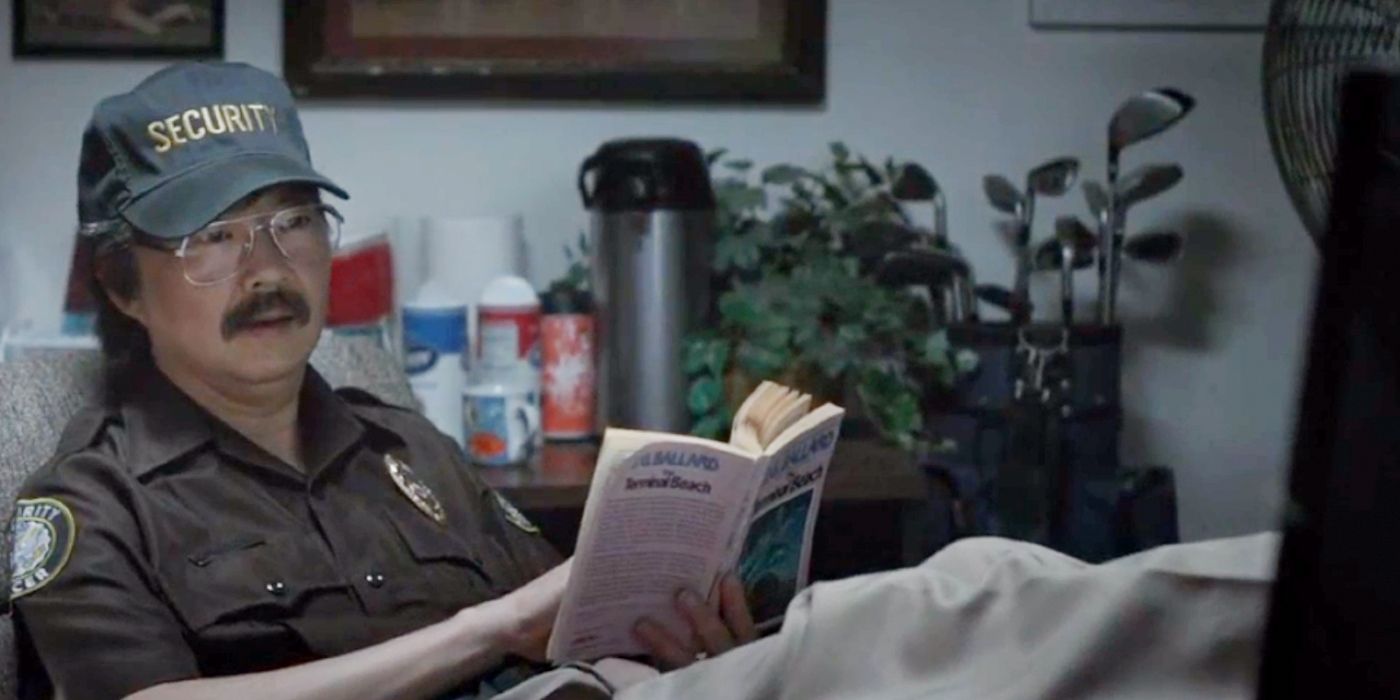 Ken Jeong dressed as a security guard sits back and reads a book