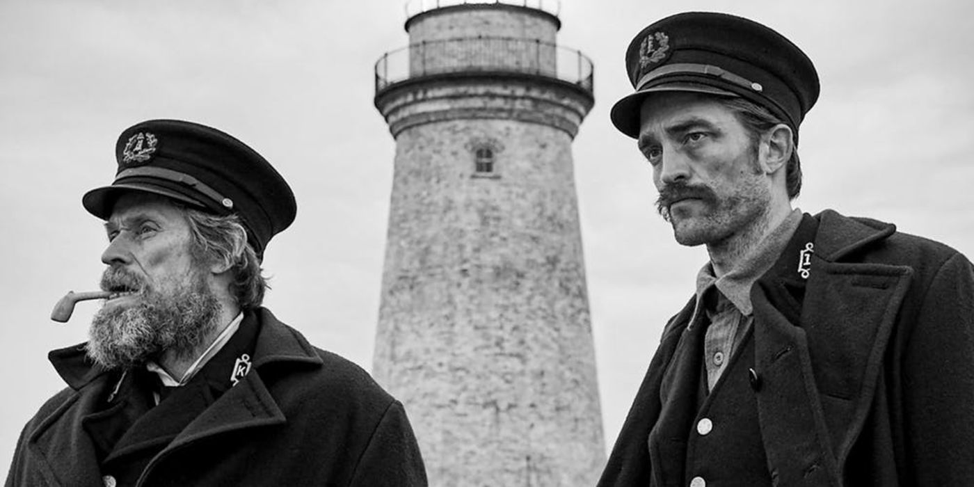 Two men stand in front of a lighthouse in The Lighthouse.