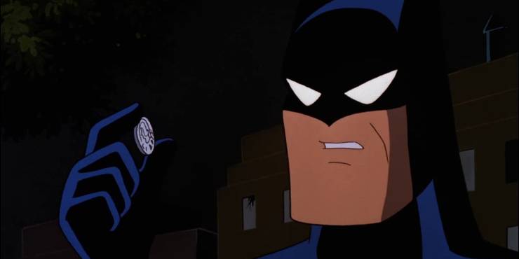 A screenshot from Batman the Animated Series. Batman wears his cowl and peers closely at a coin held between his fingers.
