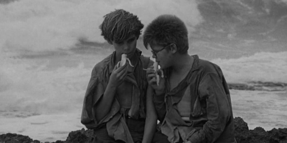 Two boys eating bananas by the shoreline in Lord of the Flies