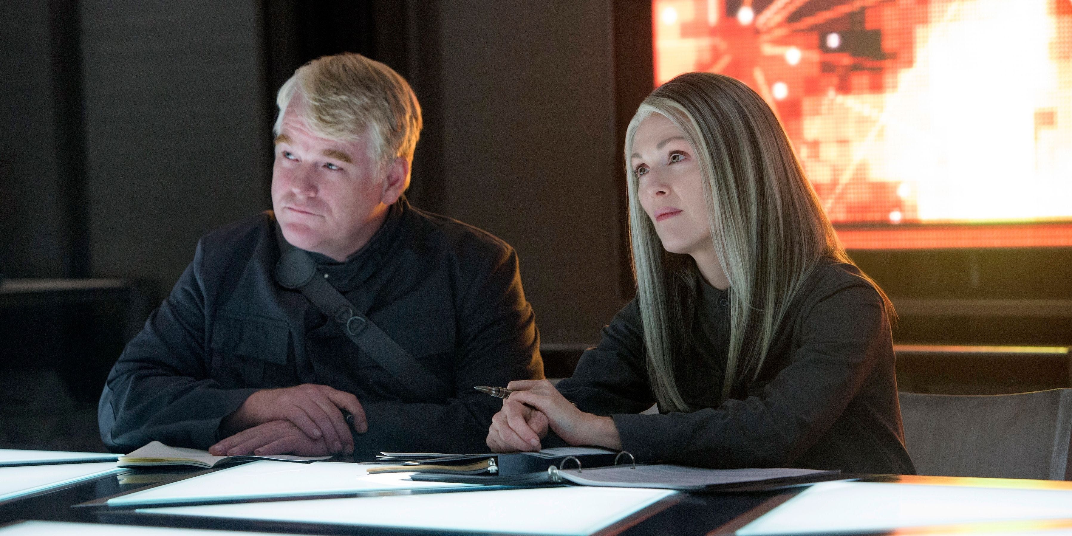 Plutarch Heavensbee and Alma Coin sitting behind a desk and looking intently in The Hunger Games Mockingjay.