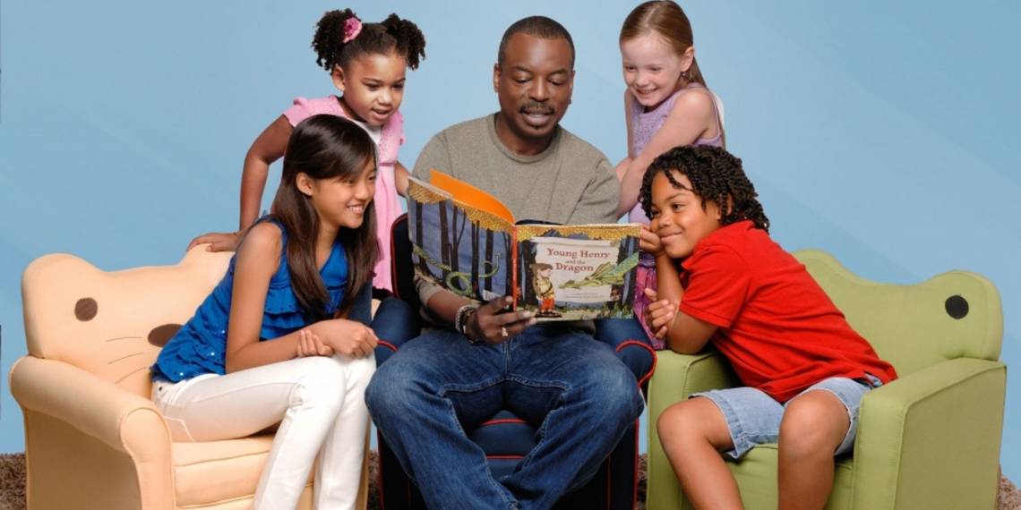 LeVar Burton reading to a group of kids. Burton holds a picture book and sits in the center while kids circle around him.