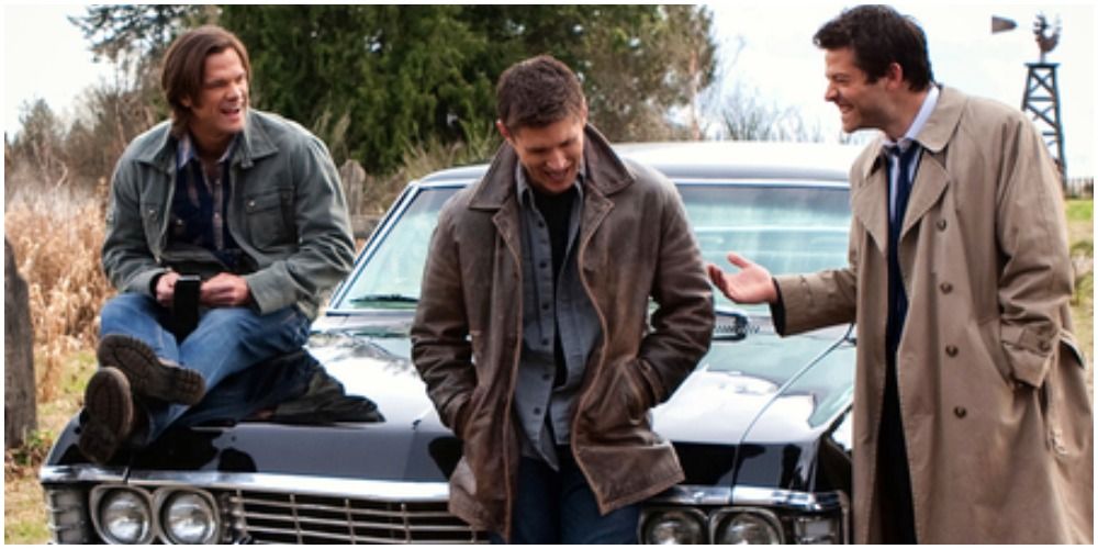 Left to right Sam sitting on hood of Impala, Dean standing hands in pockets, Castiel leaning toward Dean laughing