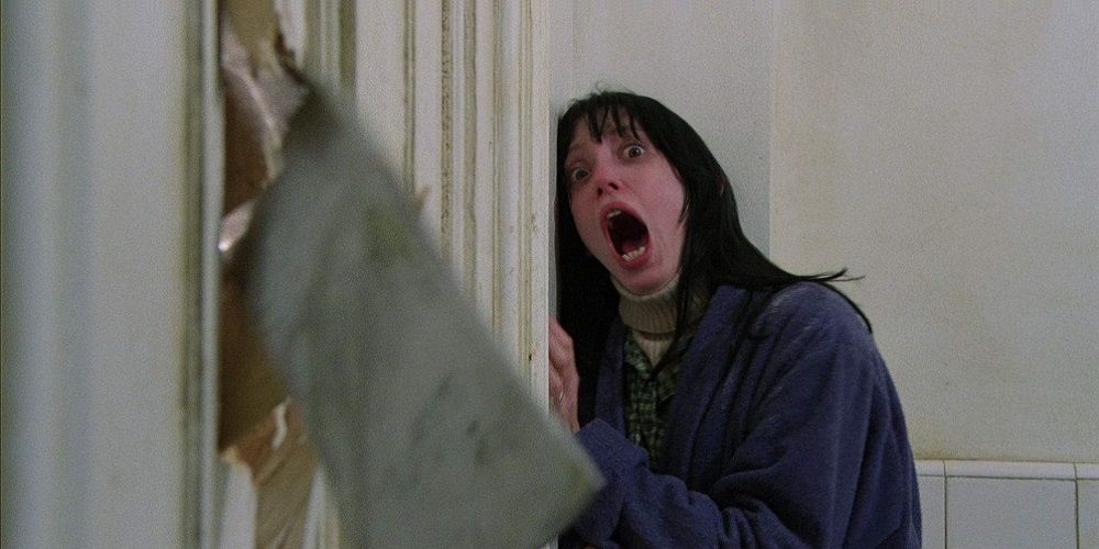 Wendy cowers in fear as Jack destroys a door with an axe in The Shining