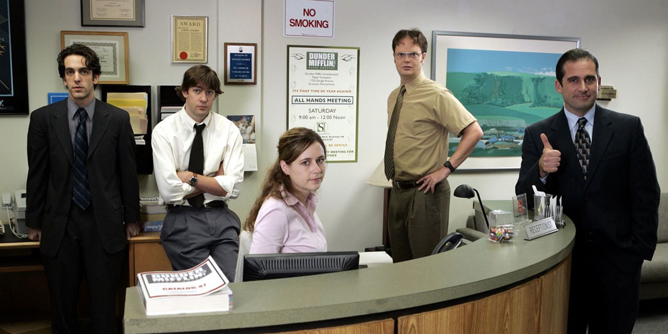 A scene from The Office with the group standing around Pam’s desk.