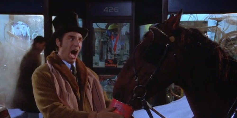 Kramer is wearing a top hat and winter coat as he feeds a horse