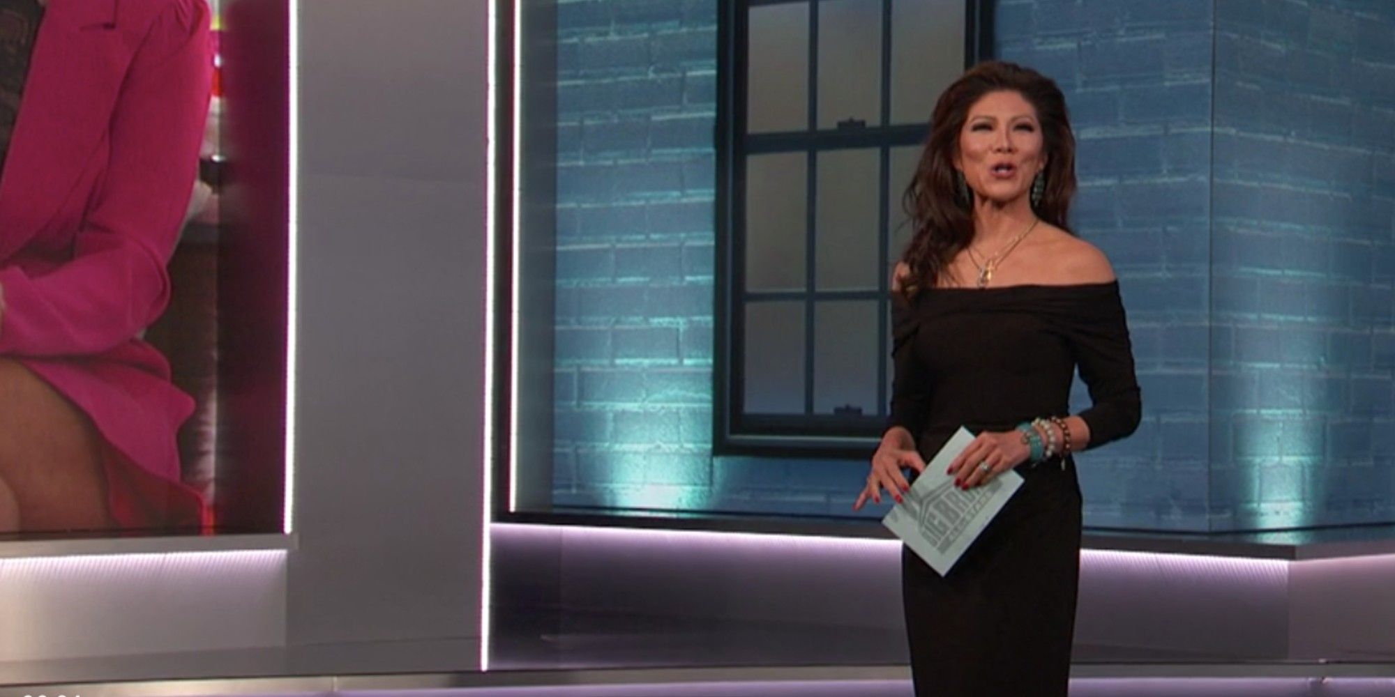 Julie Chen standing outside of the Big Brother house, holding a cue card and wearing a black off-shoulder dress.
