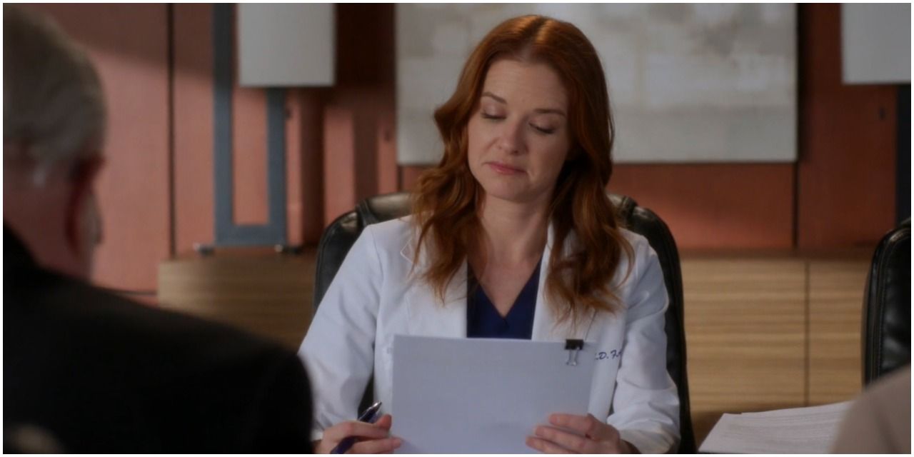 An image of April Kepner in Grey's Anatomy. She is seen to be wearing dark blue scrubs and a white lab coat. She is also seen sitting at a table reading some paperwork