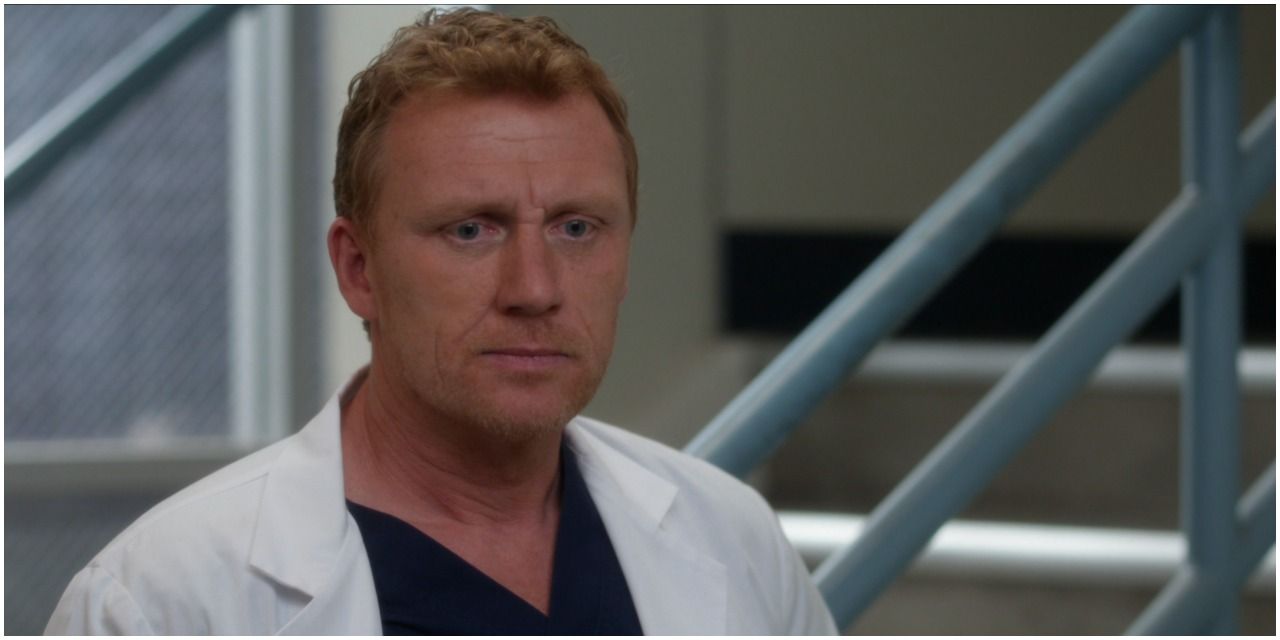 An image of Owen Hunt in Grey&rsquo;s Anatomy, wearing dark blue scrubs and a white lab coat. He appears to be standing in a stairwell as there are stairs in the background. Owen looks like he will head up them