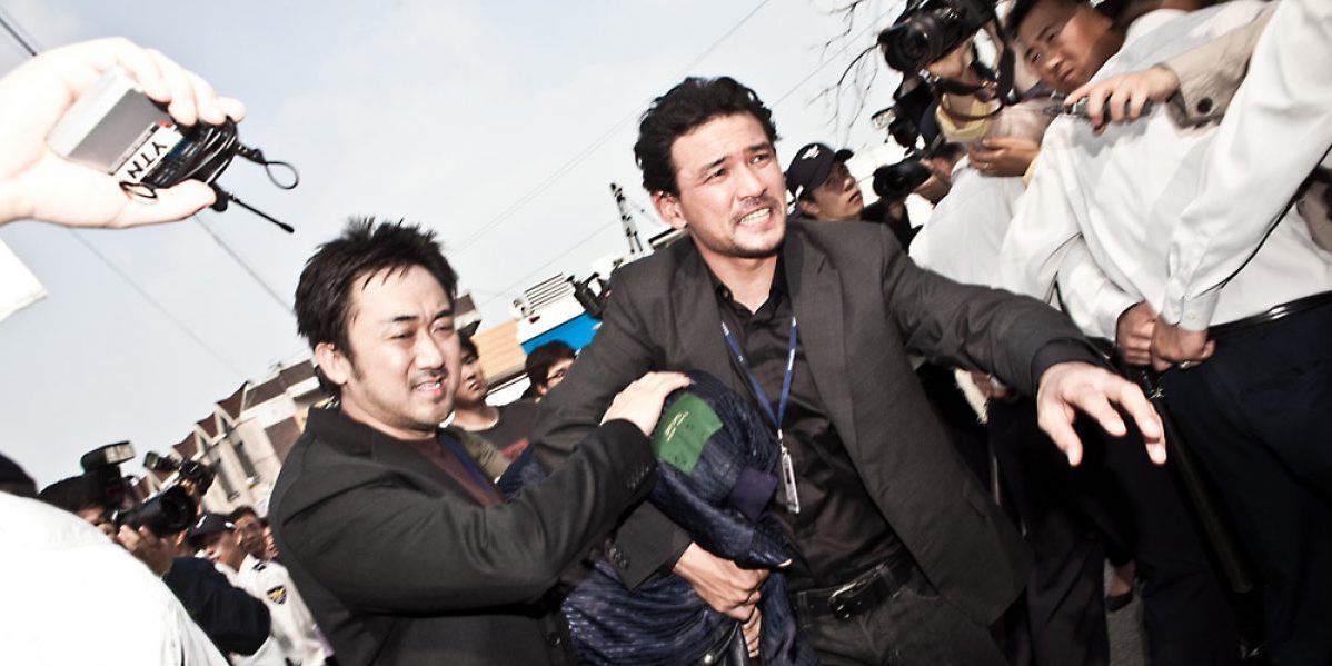 Hwang Jung-min and Don Lee rushing a suspect past a crowd with a jacket over their head in The Unjust