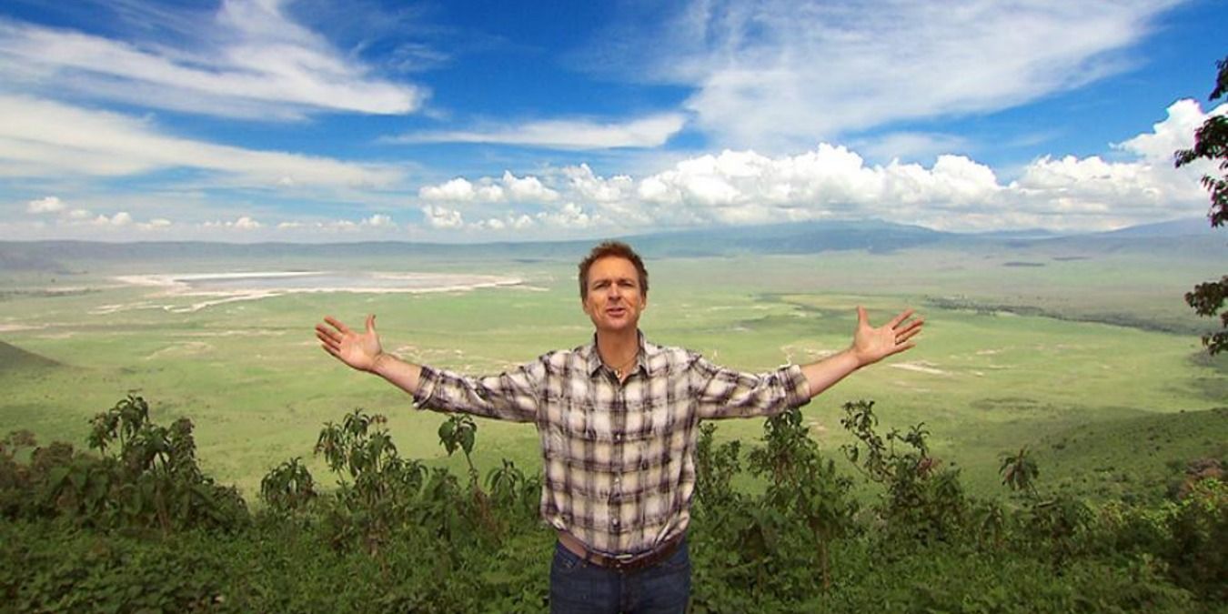 Phil Keoghan, arms wide open, against bucolic backdrop in The Amazing Race
