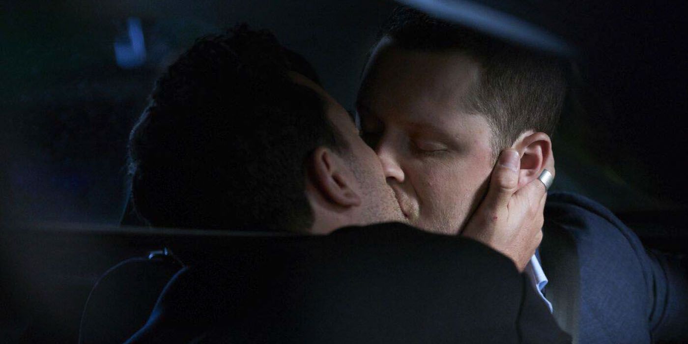 A man with dark brown hair holding the face of another man with light brown hair, kissing him. It is a close up but you can tell they are in the car, the lighter brunette is wearing a seat belt.