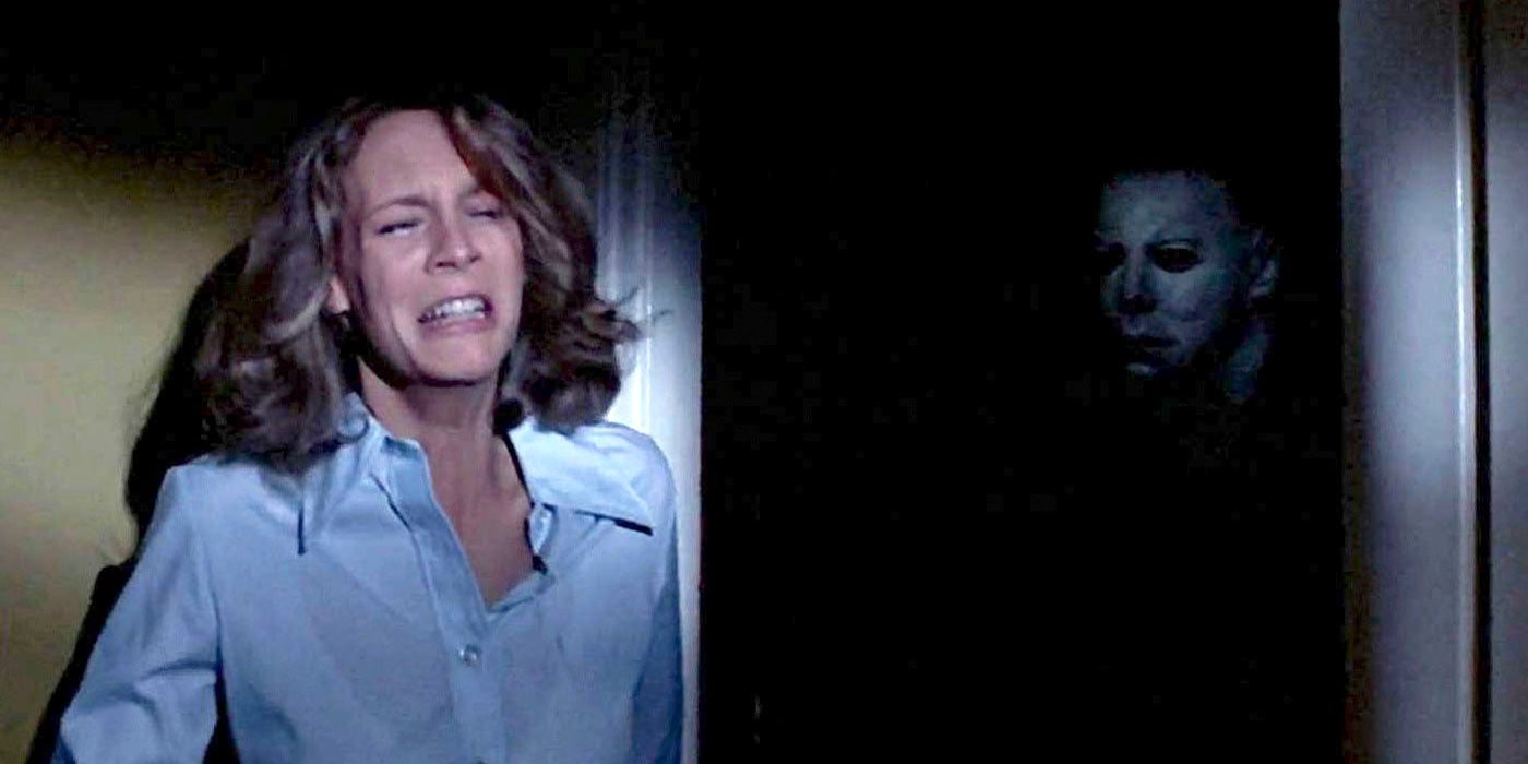 Laurie rests agains a wall while Michael Myers lurks in the darkness from Halloween