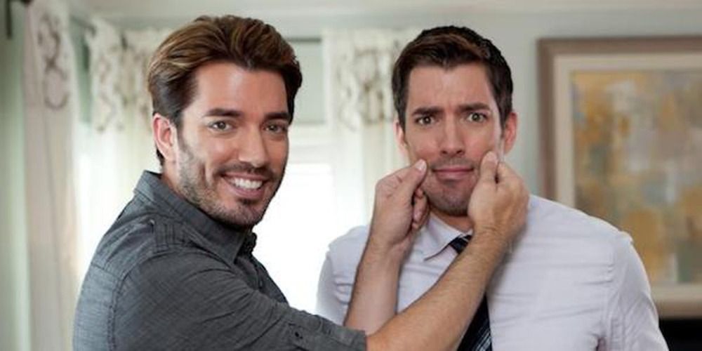 The two Property Brothers joking around with the one who does the manual labor pinching the cheeks of the one who wears and suit and tie 