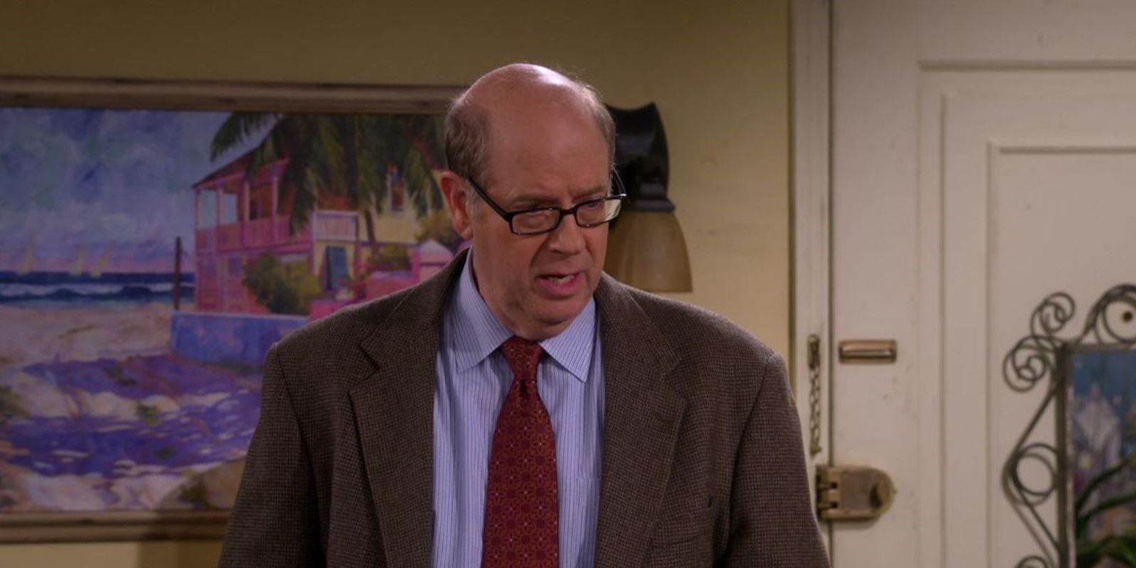 Stephen Tobolowsky in One Day at a Time standing in a room with a suit on, looking down.