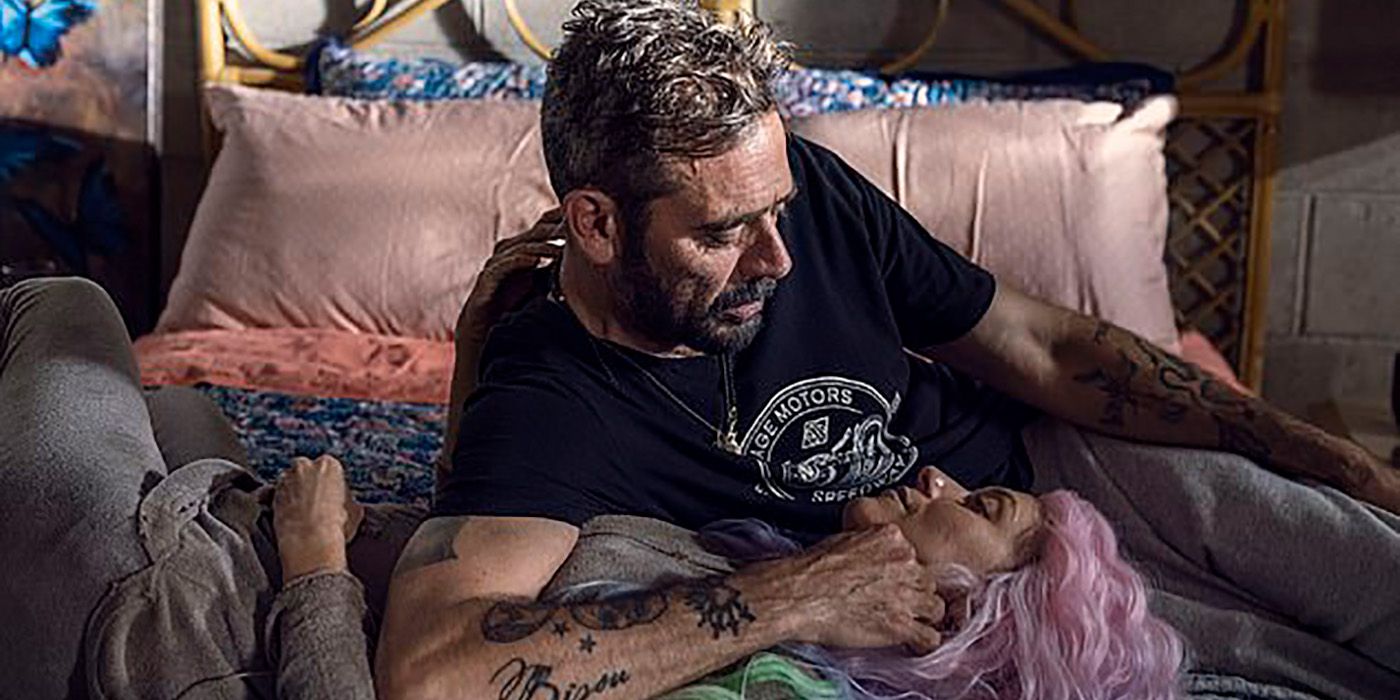 Negan lying on a bed cradling Lucille's head in his arms, her wearing a pink wig