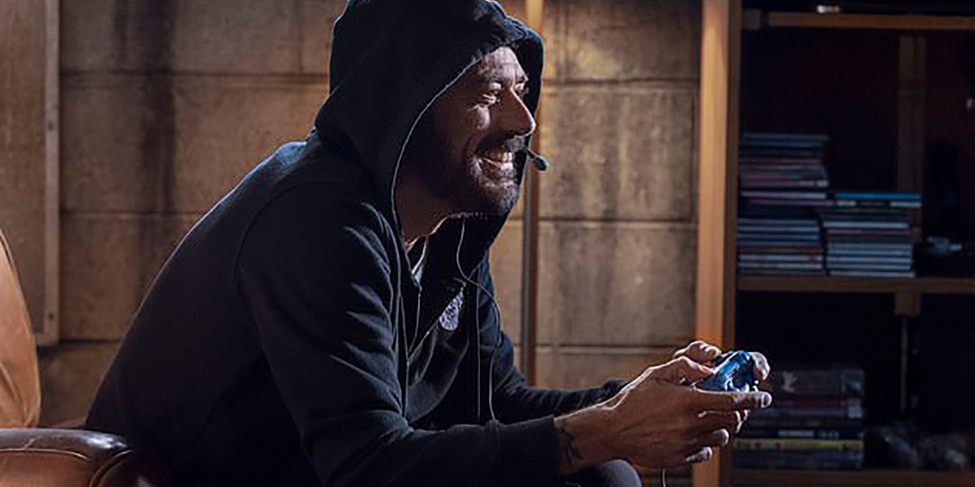 A side view of Negan wearing a black hoodie with a smile and a video game controller in his hand