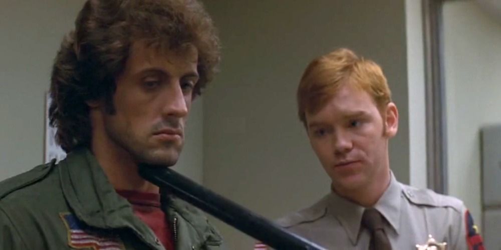 A police officer holds their baton up to Rambo's chin in First Blood