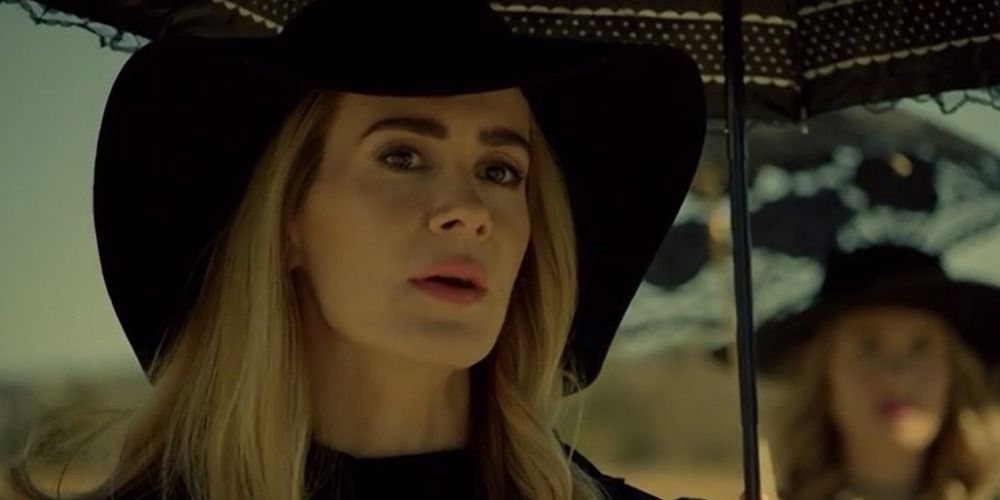 Cordelia looking at something off-screen while wearing a hat and holding an umbrella in Coven